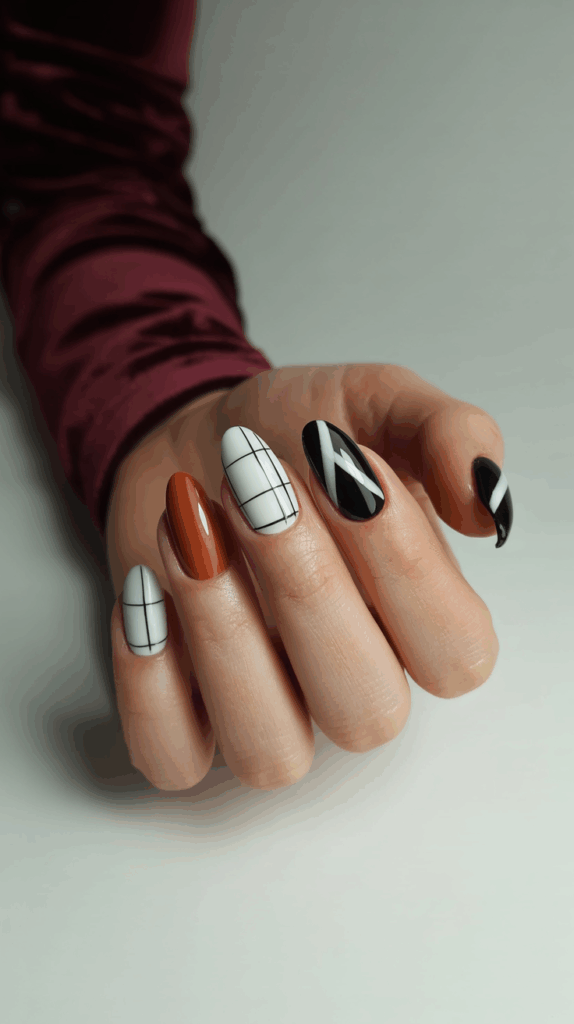Close-up of a hand with neatly manicured nails featuring a mix of white nails with black grid designs, a solid orange nail, and black nails with white diagonal stripes, set against a soft white background.