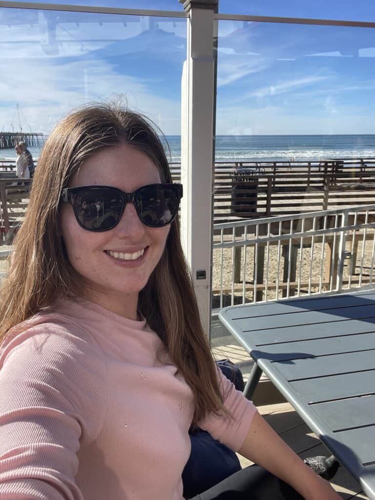 A woman wearing sunglasses and a pink top smiles while taking a selfie on an outdoor patio by the beach, with the ocean visible in the background under a clear blue sky.