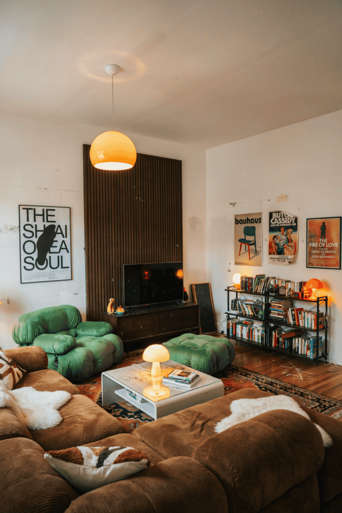 A cozy living room with a brown sectional sofa, green plush chairs, bookshelves, and posters on the walls.